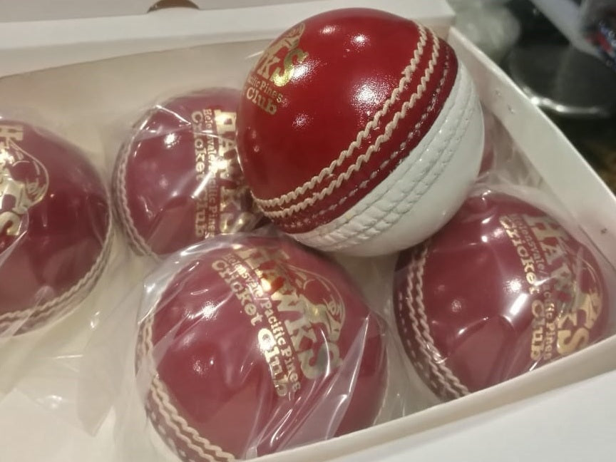 A close-up image of a Super Match 156g Dual Color cricket ball by Advance, featuring red leather with white stitching. The side of the hand-stitched ball displays a logo and text, and its full cork core and leather coating give it a new, shiny appearance against the white background.