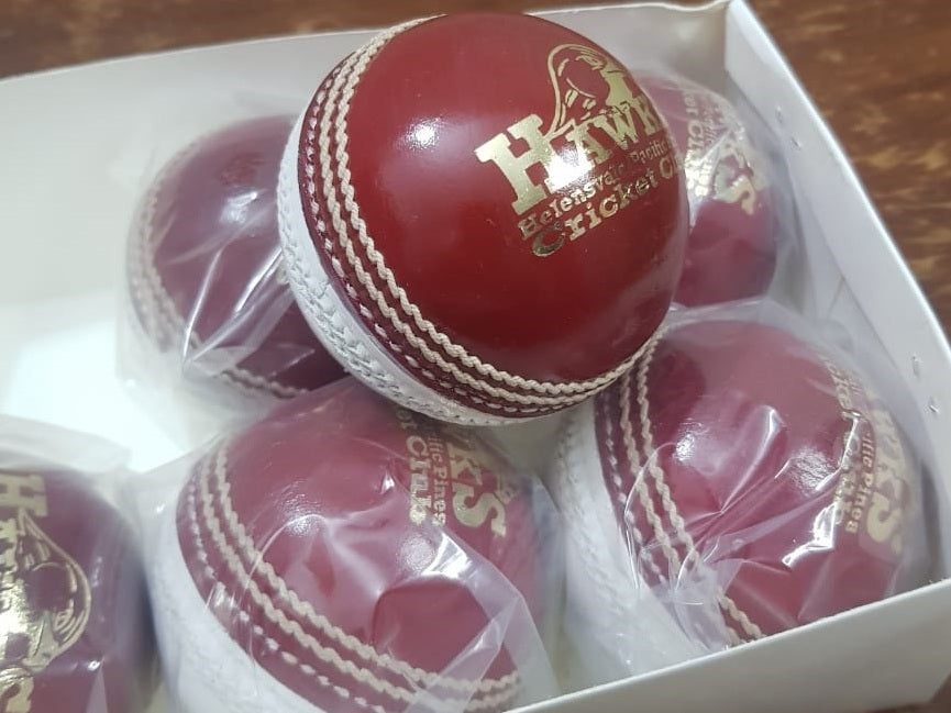 A close-up image of a Super Match 156g Dual Color cricket ball by Advance, featuring red leather with white stitching. The side of the hand-stitched ball displays a logo and text, and its full cork core and leather coating give it a new, shiny appearance against the white background.
