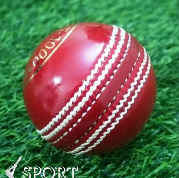 A close-up of a red leather cricket ball from the Target 1000 Super Test pack by Advance, featuring prominent white hand-stitched seams. The ball appears new and shiny with gold lettering partially visible around its circumference, displaying "STA" and "ADVAN.