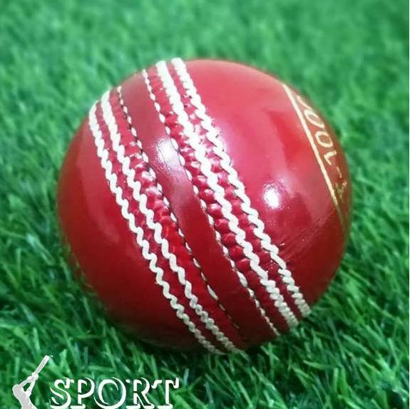 A close-up of a red leather cricket ball from the Target 1000 Super Test pack by Advance, featuring prominent white hand-stitched seams. The ball appears new and shiny with gold lettering partially visible around its circumference, displaying "STA" and "ADVAN.