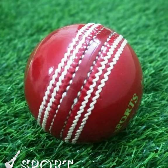 A close-up of a red leather cricket ball from the Target 1000 Super Test pack by Advance, featuring prominent white hand-stitched seams. The ball appears new and shiny with gold lettering partially visible around its circumference, displaying "STA" and "ADVAN.